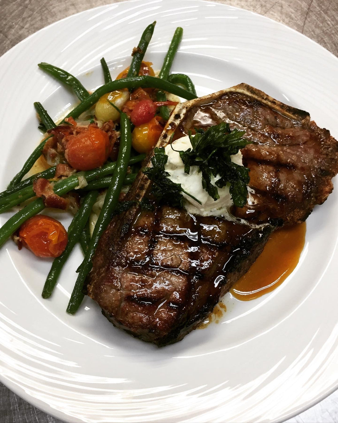 Grilled bone-in strip steak topped with herb butter and demi-glace, served with sautéed green beans, bacon, and tomatoes